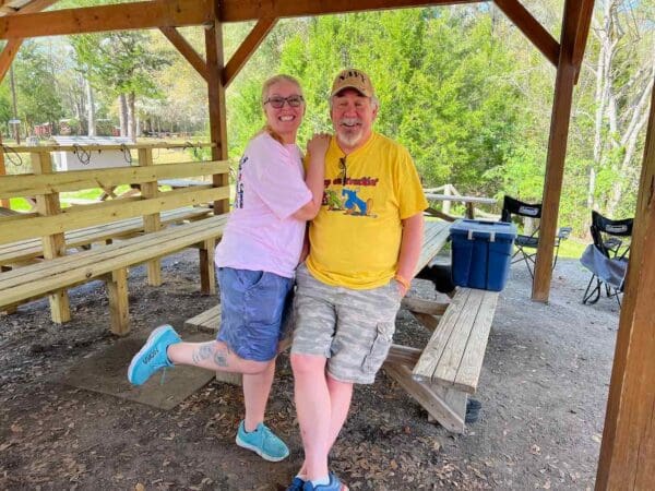 Two people posing for a picture in front of a park bench.