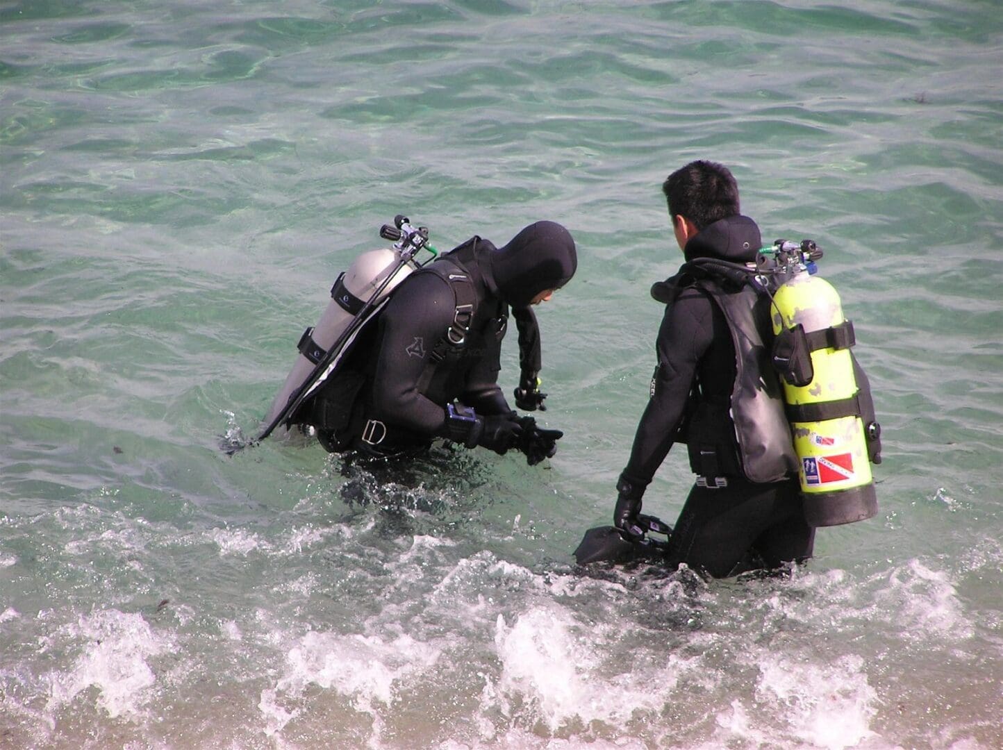 Two people in wetsuits and scuba gear are wading into the ocean.